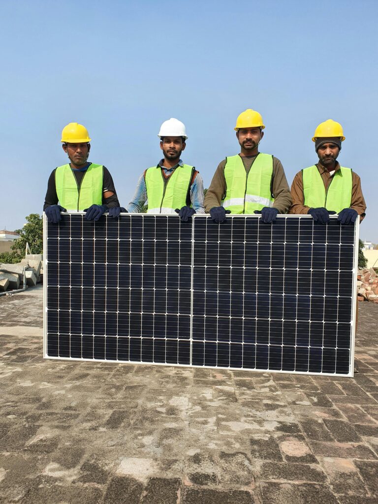 Group of construction workers holding a solar panel on a sunny day, wearing safety gear.